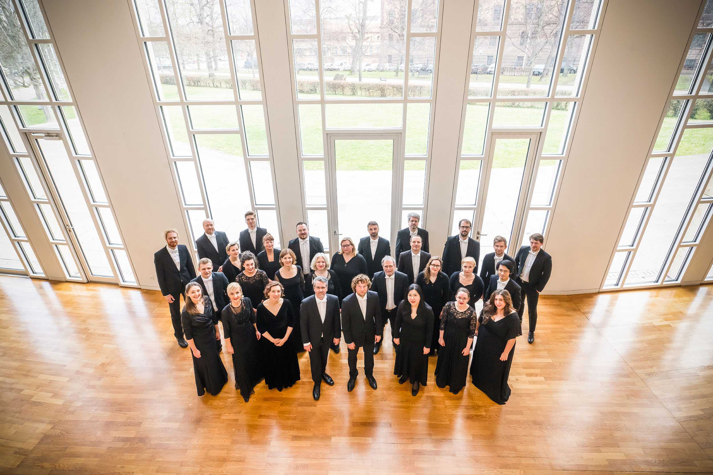 29 Chorsänger*innen der Gaechinger Cantorey stehen in einer Halle vor einer Fensterfront