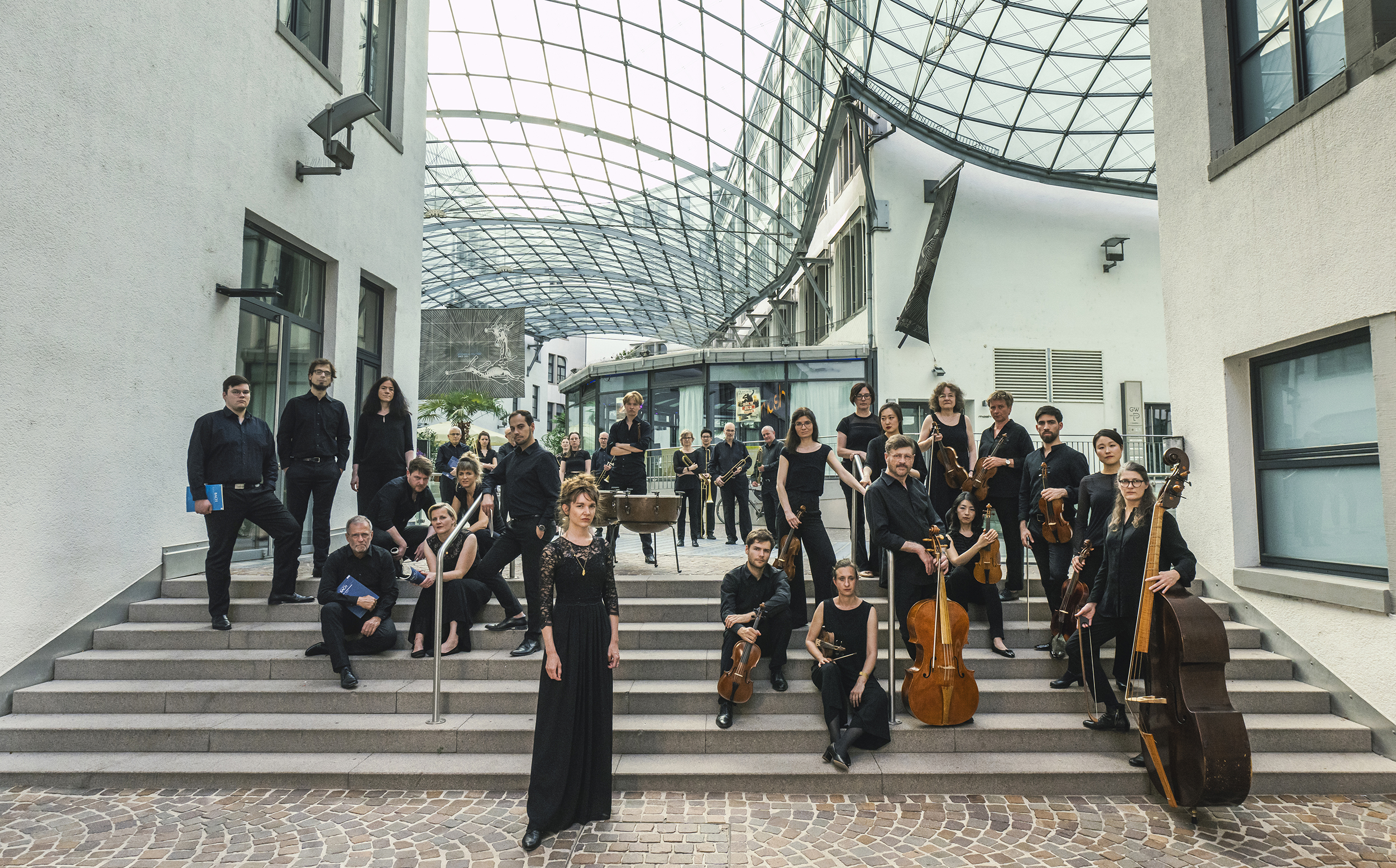 Das Bild zeigt das Orchester der Gaechinger Cantorey, das in einem modernen Treppenhaus posiert. Die Musiker stehen elegant auf den Treppenstufen und tragen formelle schwarze Kleidung. Einige halten ihre Instrumente.