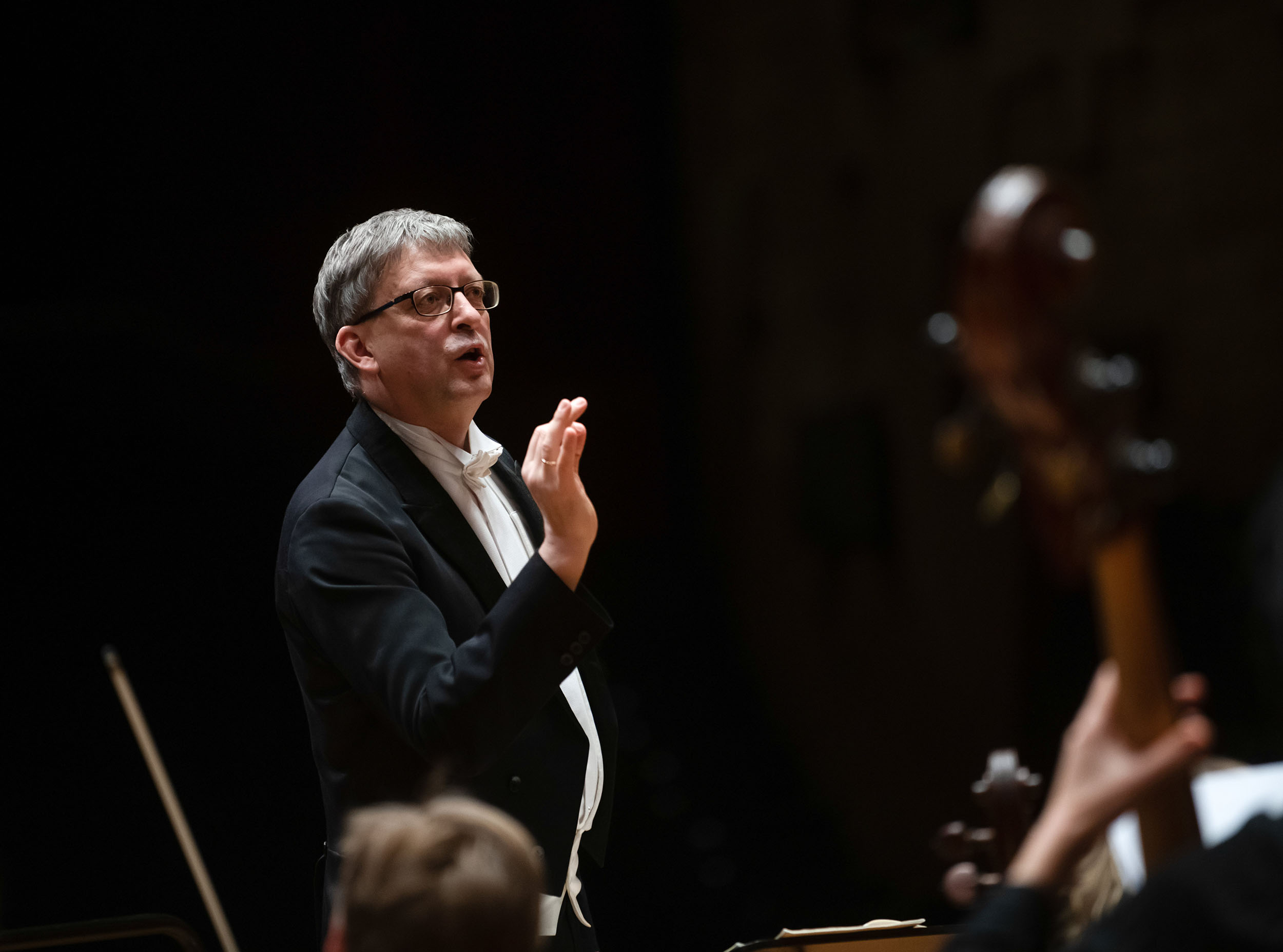 Das Bild zeigt Hans-Christoph Rademann. Er trägt einen klassischen schwarzen Anzug mit weißem Hemd und Fliege und hebt eine Hand in einer dirigierenden Geste, während er konzentriert in die Richtung des Orchesters schaut.