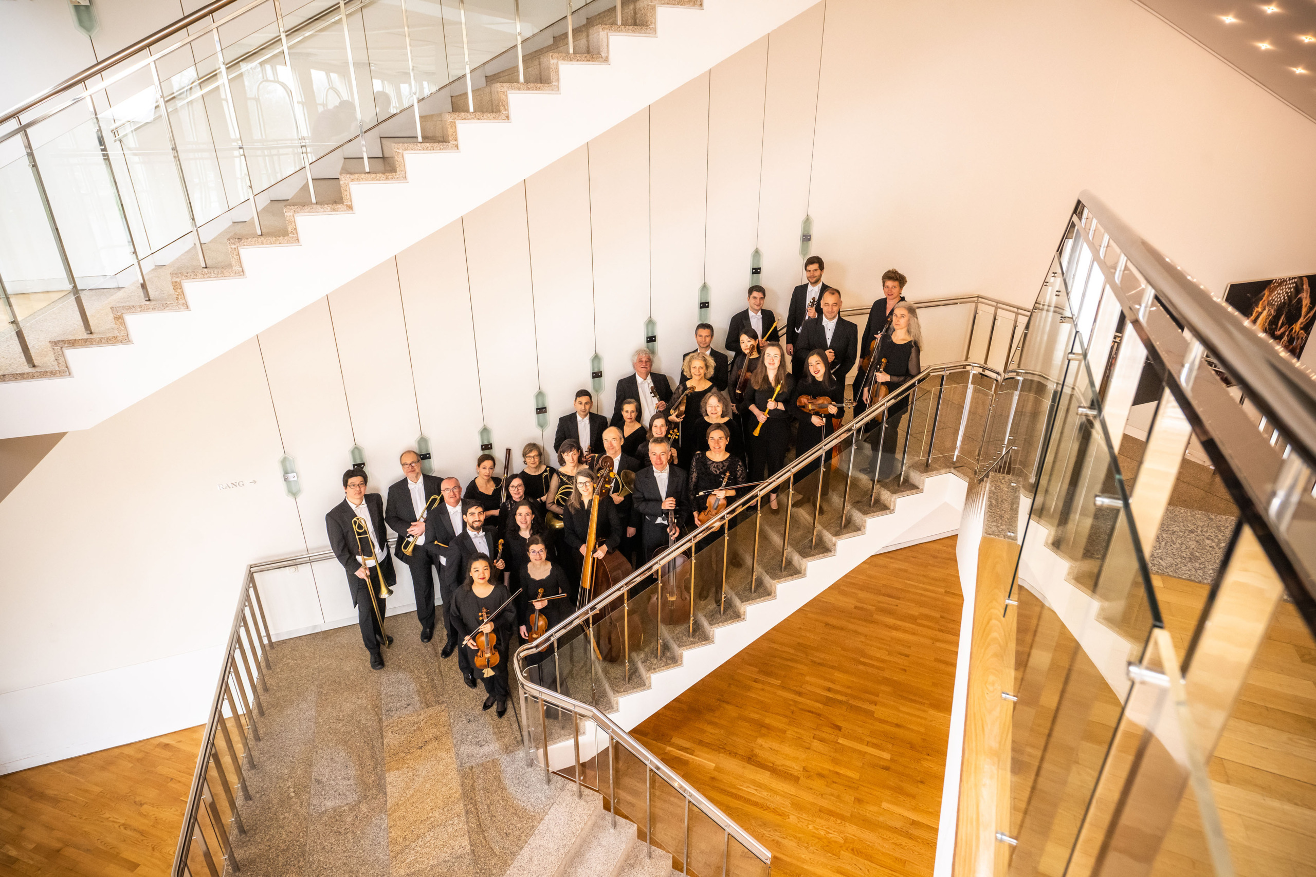 Das Bild zeigt das Orchester der Gaechinger Cantorey, das in einem modernen Treppenhaus posiert. Die Musiker stehen elegant auf den Treppenstufen und tragen formelle schwarze Kleidung. Einige halten ihre Instrumente